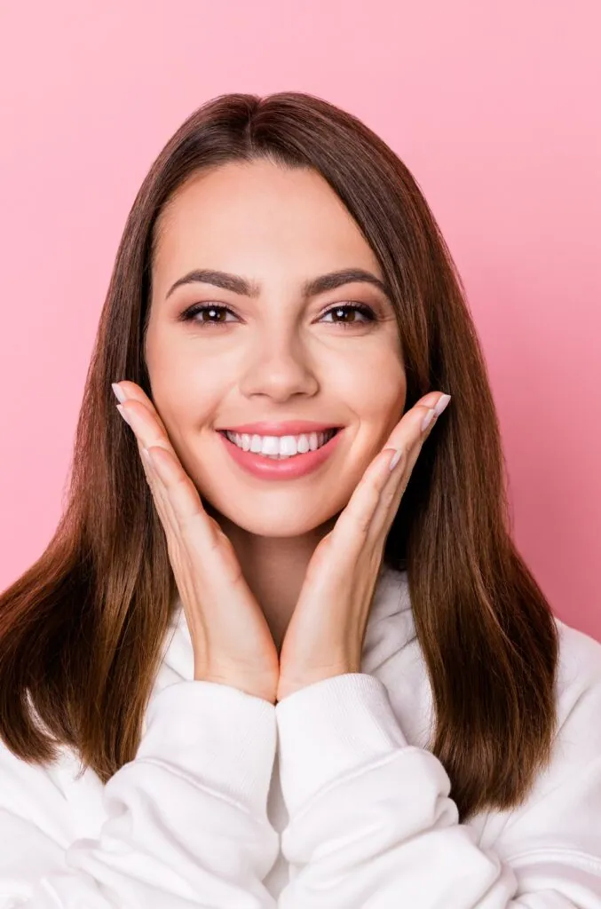 Foto de una joven atractiva y feliz que se sostiene las mejillas con las manos y muestra los dientes, aislada sobre un fondo rosado