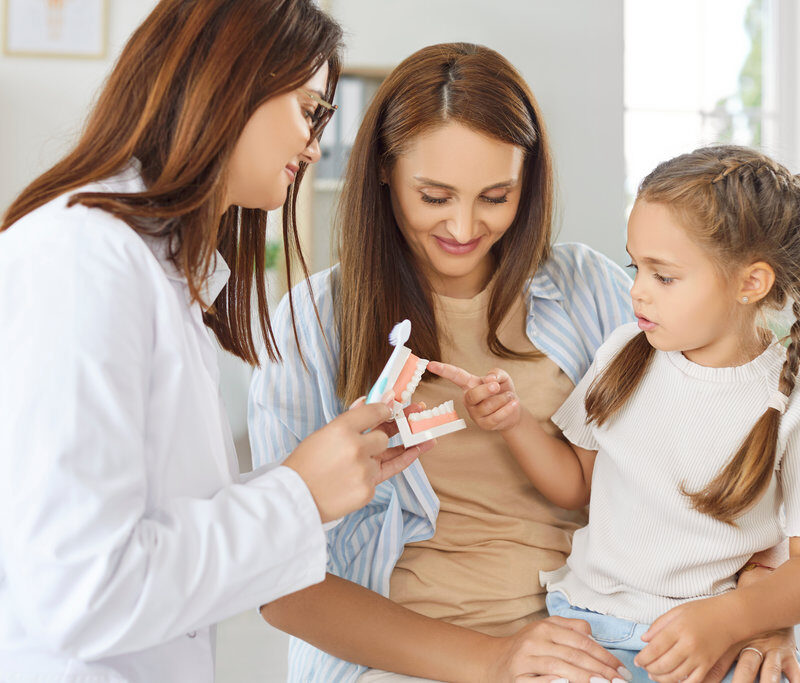 Dentist doctor or nurse demonstrates teeth cleaning techniques to a parent and child. The oral care session helps teach the child how to use a toothbrush during visit to the clinic.