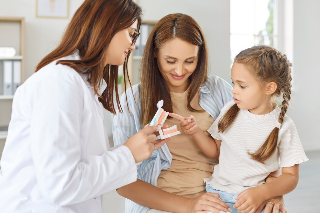 Dentist doctor or nurse demonstrates teeth cleaning techniques to a parent and child. The oral care session helps teach the child how to use a toothbrush during visit to the clinic.