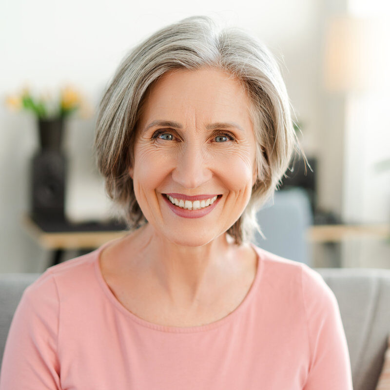Confident smiling senior woman sitting on sofa looking at camera. Portrait happy retired female with stylish gray hair, white teeth at home. Natural beauty, healthy lifestyle concept
