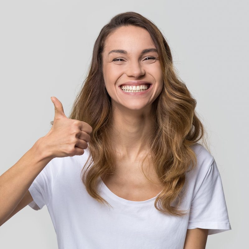 Young satisfied woman smiling with white teeth showing thumbs up looking at camera isolated on studio blank background, happy girl student like recommend dental orthodontic service concept, portrait